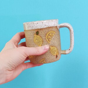 May include: A ceramic mug with a speckled brown glaze and a white rim. The mug has a handle and is decorated with yellow lemons and green leaves.
