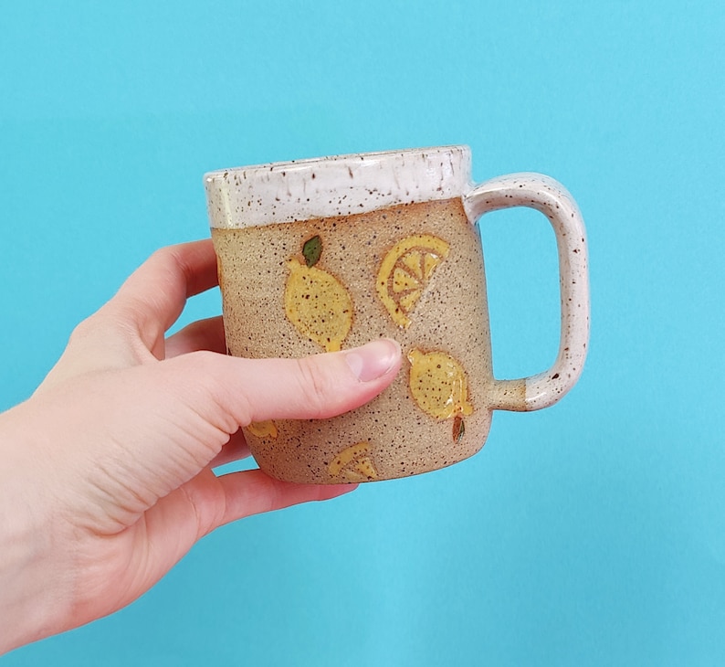 May include: A ceramic mug with a speckled brown glaze and a white rim. The mug has a handle and is decorated with yellow lemons and green leaves.