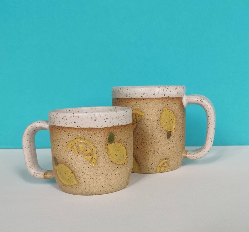 May include: Two ceramic mugs with a speckled glaze and a lemon design. The larger mug has a handle on the right side and the smaller mug has a handle on the left side.