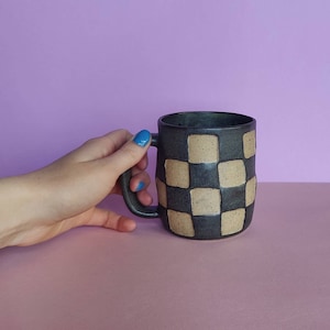 Puede incluir: Una taza de cer&aacute;mica negra con un patr&oacute;n de cuadros en beige y negro. La taza tiene un asa redondeada.