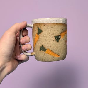 Op de afbeelding: Een gespikkelde keramische mok met een beige basis en een witte rand. De mok is versierd met oranje wortelillustraties met groene toppen. De mok heeft een gebogen handvat en wordt tegen een paarse achtergrond gehouden.