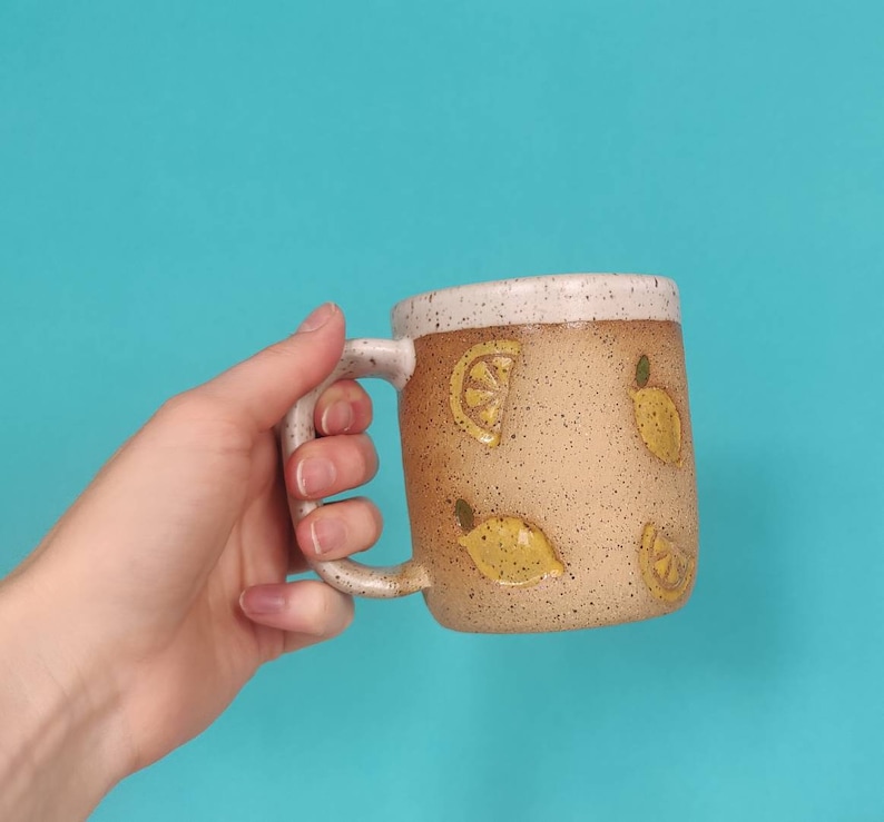 May include: A beige ceramic mug with a white rim and a speckled finish. The mug is decorated with yellow lemon slices and whole lemons.