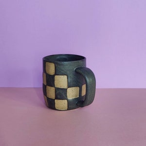 Puede incluir: Una taza de cer&aacute;mica negra con un patr&oacute;n de cuadros en color marr&oacute;n claro y negro. La taza tiene un cuerpo redondeado y un asa.