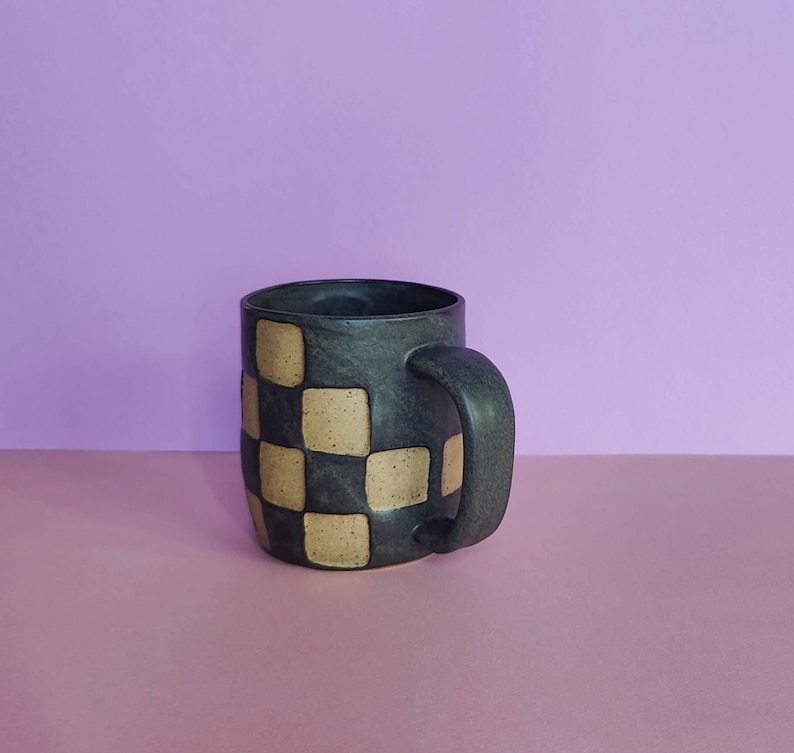 Puede incluir: Una taza de cer&aacute;mica negra con un patr&oacute;n de cuadros en color marr&oacute;n claro y negro. La taza tiene un cuerpo redondeado y un asa.