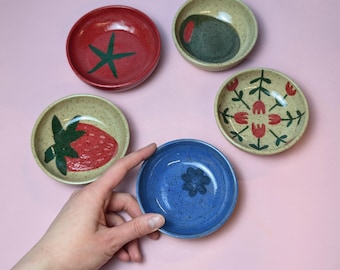 May include: Five small ceramic bowls with different designs. The bowls are in various colors, including red, blue, green, and beige. The designs include a star, a strawberry, a flower, and a geometric pattern.