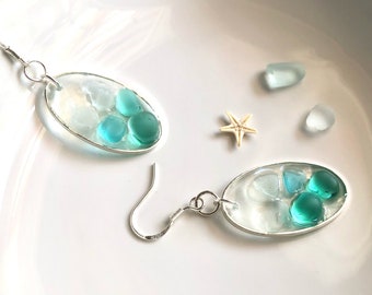 May include: A pair of silver earrings with oval-shaped resin pendants. Each pendant features a clear resin base with embedded blue and green sea glass pieces.