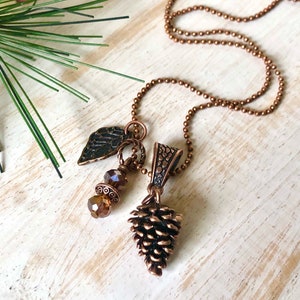 May include: A copper-coloured necklace with a pine cone pendant, a leaf charm, and brown crystal beads. The necklace has a ball chain and is set against a rustic, light-coloured wooden surface with a sprig of pine needles.