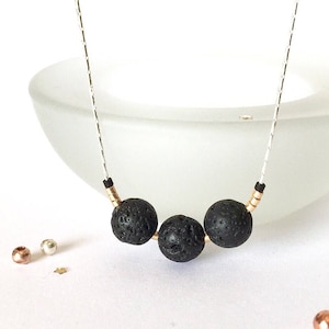 May include: A necklace with three black lava stone beads on a silver chain. The beads are separated by gold-coloured spacers. The necklace is set against a white background with a blurred glass bowl. The necklace is a stylish accessory.