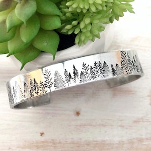 May include: A silver cuff bracelet featuring a black forest silhouette design. The forest scene, with trees and ferns, wraps around the bracelet. The open-ended bracelet is displayed on a light-coloured wooden surface.