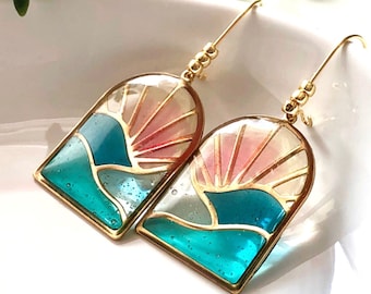 May include: A pair of gold earrings with a stained glass design. The earrings feature a pink and blue sunset with a gold frame.