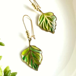 May include: A pair of gold earrings with green leaf charms. The leaves are detailed with gold veins and have a green enamel finish.