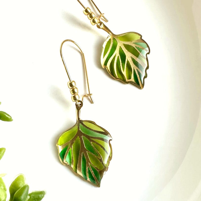 May include: A pair of gold earrings with green leaf charms. The leaves are detailed with gold veins and have a green enamel finish.
