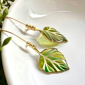 May include: Gold leaf earrings with green and gold enamel. The leaves are detailed with gold veins and have a glossy finish.