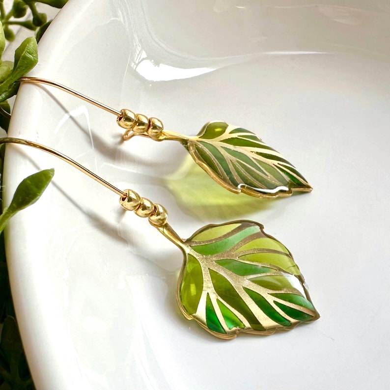 May include: Gold leaf earrings with green and gold enamel. The leaves are detailed with gold veins and have a glossy finish.