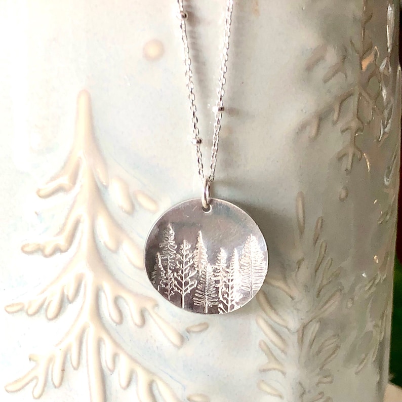 May include: A silver necklace with a round pendant featuring an embossed design of a forest with trees. The necklace has a delicate chain with small beads.