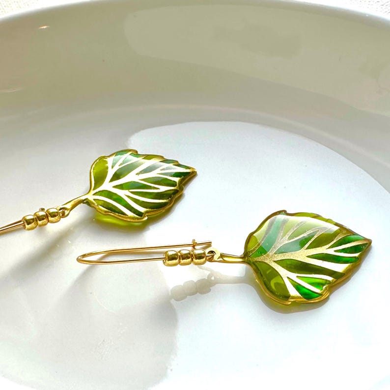 May include: A pair of gold leaf earrings with green and gold accents. The leaves are detailed with a delicate vein pattern.