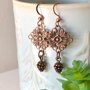 May include: A pair of copper earrings featuring a filigree design and a small pine cone charm dangling from each earring.