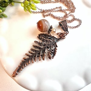 May include: A copper-toned necklace with a fern-shaped pendant. The necklace includes a chain, a round, striped stone, and a small, gold-coloured bead. The pendant features detailed leaf patterns, creating a nature-inspired accessory.