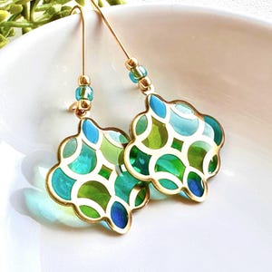 May include: Gold-tone drop earrings with a quatrefoil design. The earrings have a mosaic of green, teal, and blue glass pieces, outlined in gold. Small blue and gold beads are at the top of the earrings.