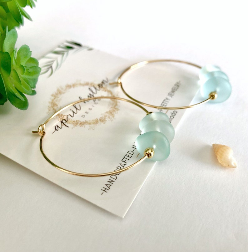 May include: Gold hoop earrings with three light blue glass beads. The earrings are handmade and feature a simple, elegant design.