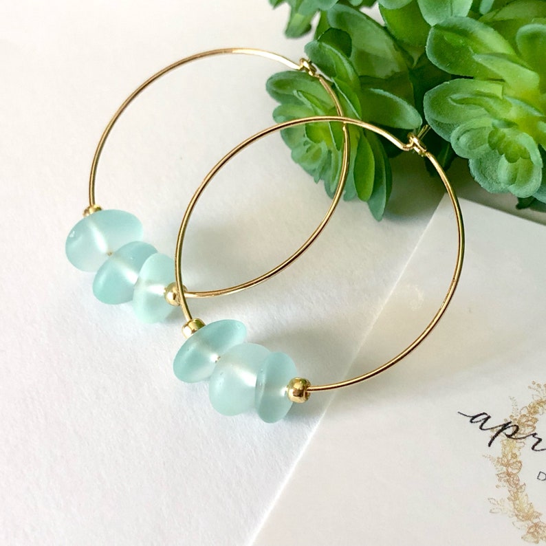 May include: Gold hoop earrings with light blue sea glass beads. The earrings are on a white surface with a card that says "apr".