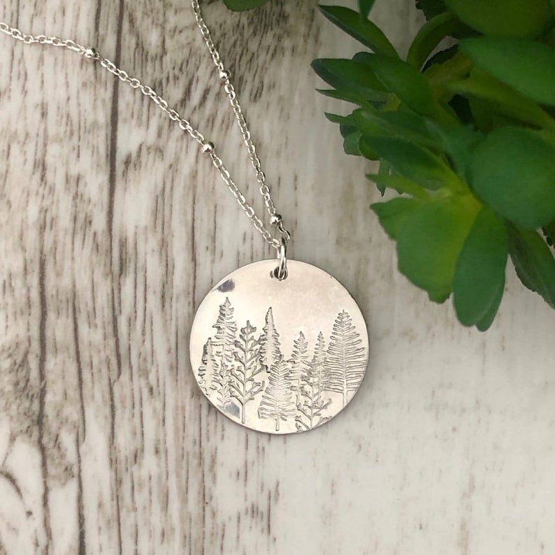 May include: A silver necklace with a round pendant featuring an etched design of a forest with trees.