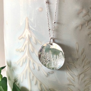 May include: A silver necklace with a round pendant featuring an embossed design of a forest with trees. The necklace has a delicate chain with small beads.