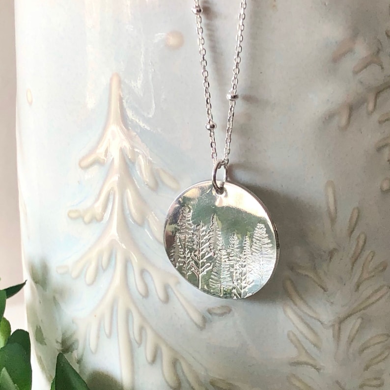 May include: A silver necklace with a round pendant featuring an embossed design of a forest with trees. The necklace has a delicate chain with small beads.