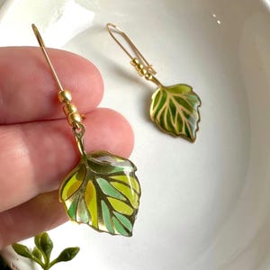 May include: Gold tone earrings with green and yellow enamel leaf charms. The leaves have a detailed vein pattern and are attached to a gold tone chain with small gold beads.