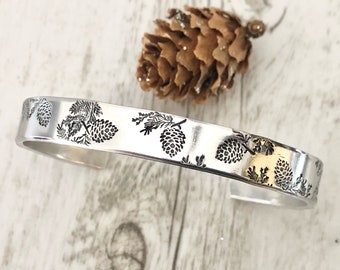 May include: A silver bracelet with a repeating pattern of pine cones and branches. The bracelet is made of metal and has a smooth, polished finish.
