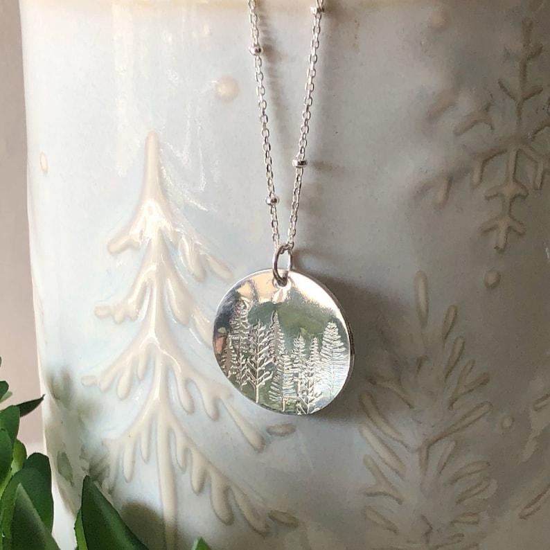 May include: A silver necklace with a round pendant featuring an embossed design of a forest with trees. The necklace is on a white background.