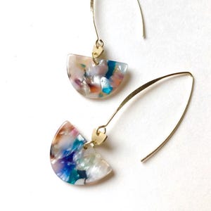 Peut inclure: Une paire de boucles d'oreilles en or avec un pendentif en forme de demi-cercle. Les pendentifs sont en résine blanche avec un motif marbré coloré.