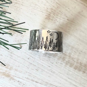 May include: A silver ring featuring a design of a forest scene with trees, a crescent moon, and stars.