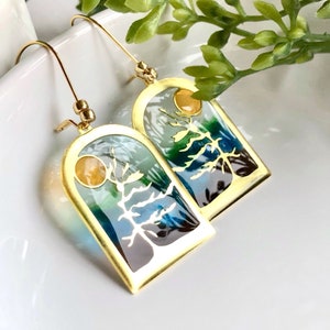 May include: A pair of gold-coloured earrings with a nature-themed design. Each earring has a rectangular frame with a tree silhouette and a colourful landscape in blue, green, and white. A small, round, yellow accent is at the top.