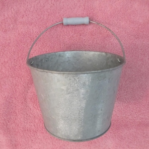 May include: A galvanized metal bucket with a gray plastic handle. The bucket has a rounded shape and a silver metal handle that arches over the top. The bucket is sitting on a pink surface.