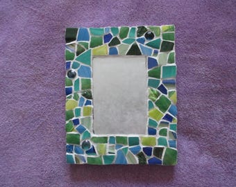 May include: A rectangular mosaic picture frame with a central opening. The frame is composed of irregularly shaped tiles in shades of green, blue, and white. The frame is approximately 6 inches tall and 4 inches wide.