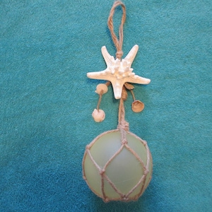 May include: A green glass fishing float with a white starfish and seashells hanging from a twine rope. The float is surrounded by a brown twine net.