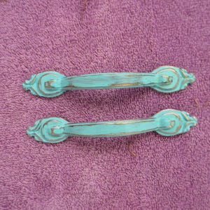 May include: Two turquoise cabinet pulls with a distressed finish. The pulls have a decorative design on each end and a curved handle. The hardware is set against a purple background.