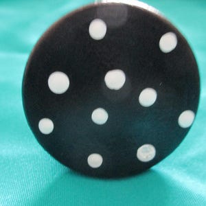 May include: A black, circular object with white polka dots. The object is set against a teal background. The polka dots are of varying sizes and are evenly spaced across the surface.