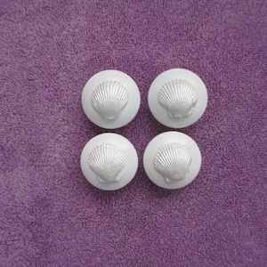 May include: Four white cabinet knobs with a seashell design. The knobs are round and have a raised, detailed seashell in the center. The background is a textured purple fabric.