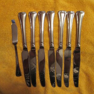May include: A set of eight silver-toned dinner knives with a polished finish. The knives have a rounded handle and a pointed blade.