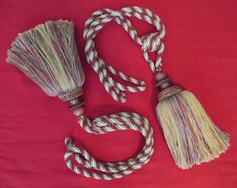 May include: Two decorative tassels with brown wooden beads and a multi-colored braided cord. The tassels are made of a soft, woven fabric in shades of brown, beige, and red.