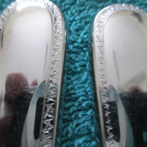 May include: Two silver-plated spoons with intricate etched designs on the handles. The spoons are lying on a teal-colored surface.