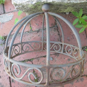 May include: A weathered, gray metal plant holder with a domed top and decorative scrollwork. The circular base and arched supports create a cage-like structure. The plant holder is resting on a brick surface.