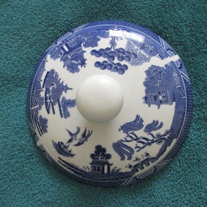 May include: A round, blue and white ceramic lid with a white knob. The lid features a Willow pattern design with scenes of pagodas, birds, and trees. The lid is likely for a teapot or serving dish.