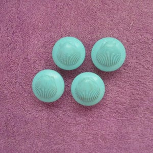May include: Four round, light blue cabinet knobs with a seashell design. The knobs are arranged on a textured, purple surface. Each knob features a raised seashell pattern in the center.