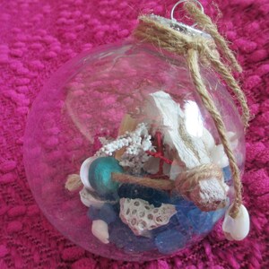 May include: A clear glass ornament filled with various sea-themed items, including seashells, coral, and blue glass beads. The ornament is tied with a brown twine bow.