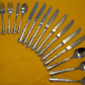 May include: A collection of stainless steel silverware, including forks, knives, spoons, and a butter knife, arranged on a yellow surface. The silverware has a decorative handle design.