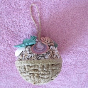 May include: A brown, woven, spherical ornament with a loop for hanging. The ornament is filled with various seashells, including a large pink shell, a smaller brown and white shell, and a green seashell.