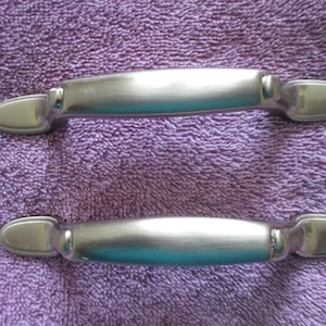 May include: Two brushed nickel cabinet door pulls with a rounded rectangular shape. The pulls have a smooth, polished finish.